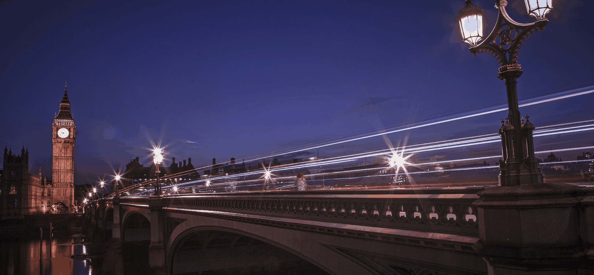 Night cityscape with an illuminated clock tower, bridge, and ornate lamppost, long-exposure light trails streaking across the scene in deep blue and warm amber tones